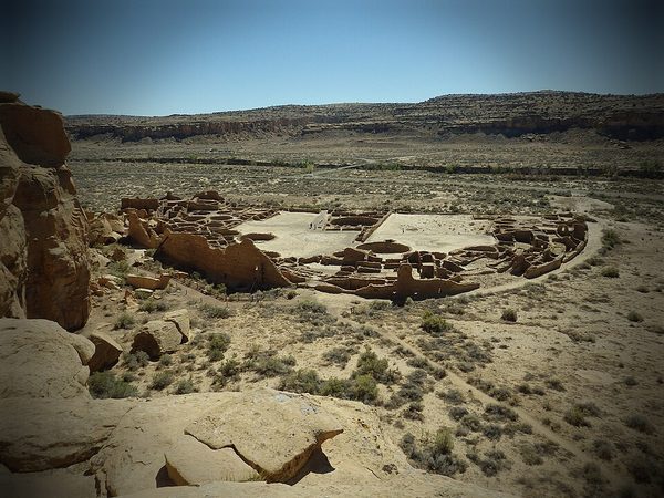 A large D-shaped stone ruin of many rooms and round kivas on a desert canyon floor