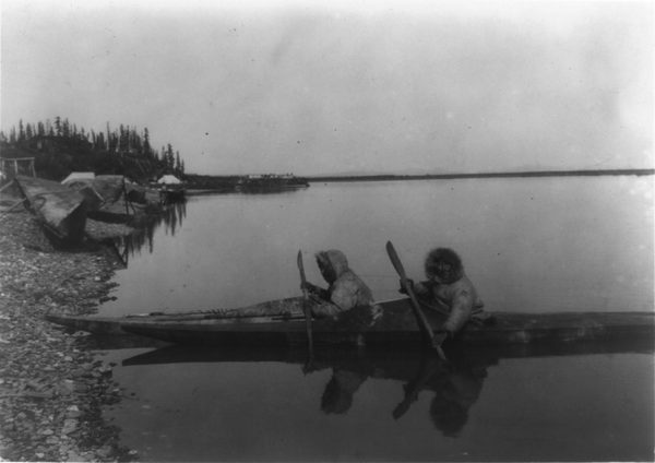 Two men in hooded parkas in kayaks at the edge of a still shore