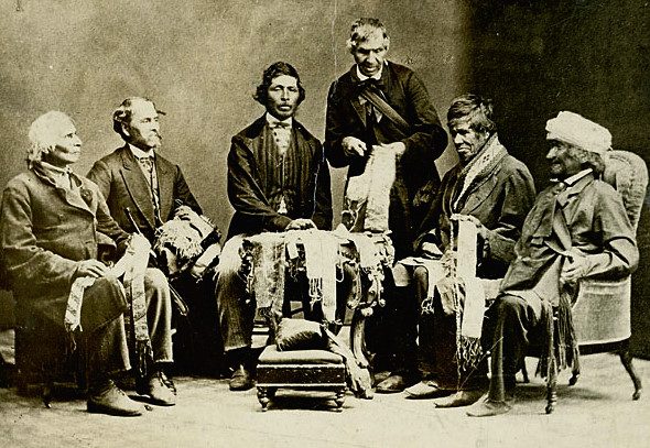 Six seated men holding wampum belts across their laps in a studio portrait